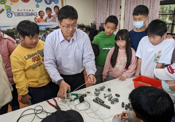 教室內，一位成年講師穿著白襯衫，向圍成一圈的小學生展示並講解各種電子零件和機械組件。學生們認真觀看桌上的教學用具，參與科技教育活動。