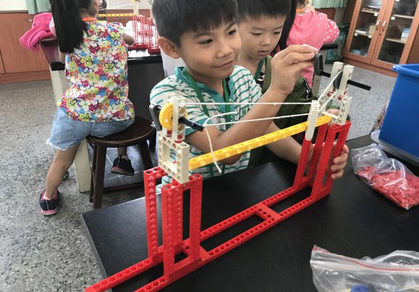 教室內，兩名小男孩正專注地組裝紅色和黃色的積木結構，背景有其他兒童在進行STEM活動。這是一堂動手建構課程，培養孩子的空間概念和問題解決能力。