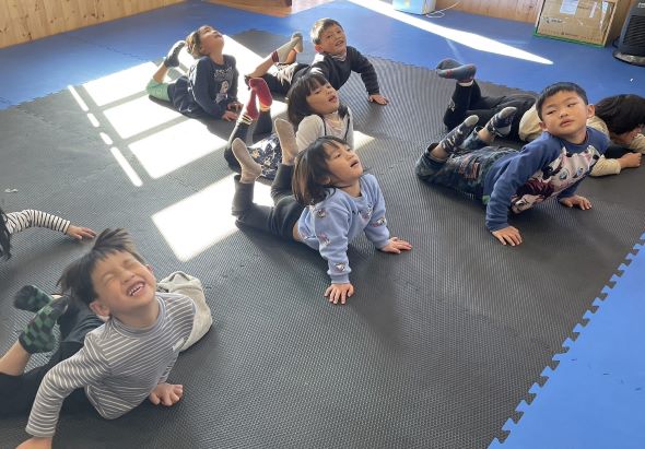 一群幼童在鋪有藍色邊框的灰色墊子上進行室內活動。孩子們以各種姿勢坐著或躺著，包括有人玩耍、互動或休息。陽光透過窗戶灑落在地面上，室內環境明亮通風。