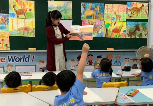 小學教室內，女教師站在講台前展示圖書，向坐在座位上舉手提問的學生們講授。黑板後方貼滿學生繪製的彩色動物畫作。教室佈置溫馨，展現互動式教學情景。