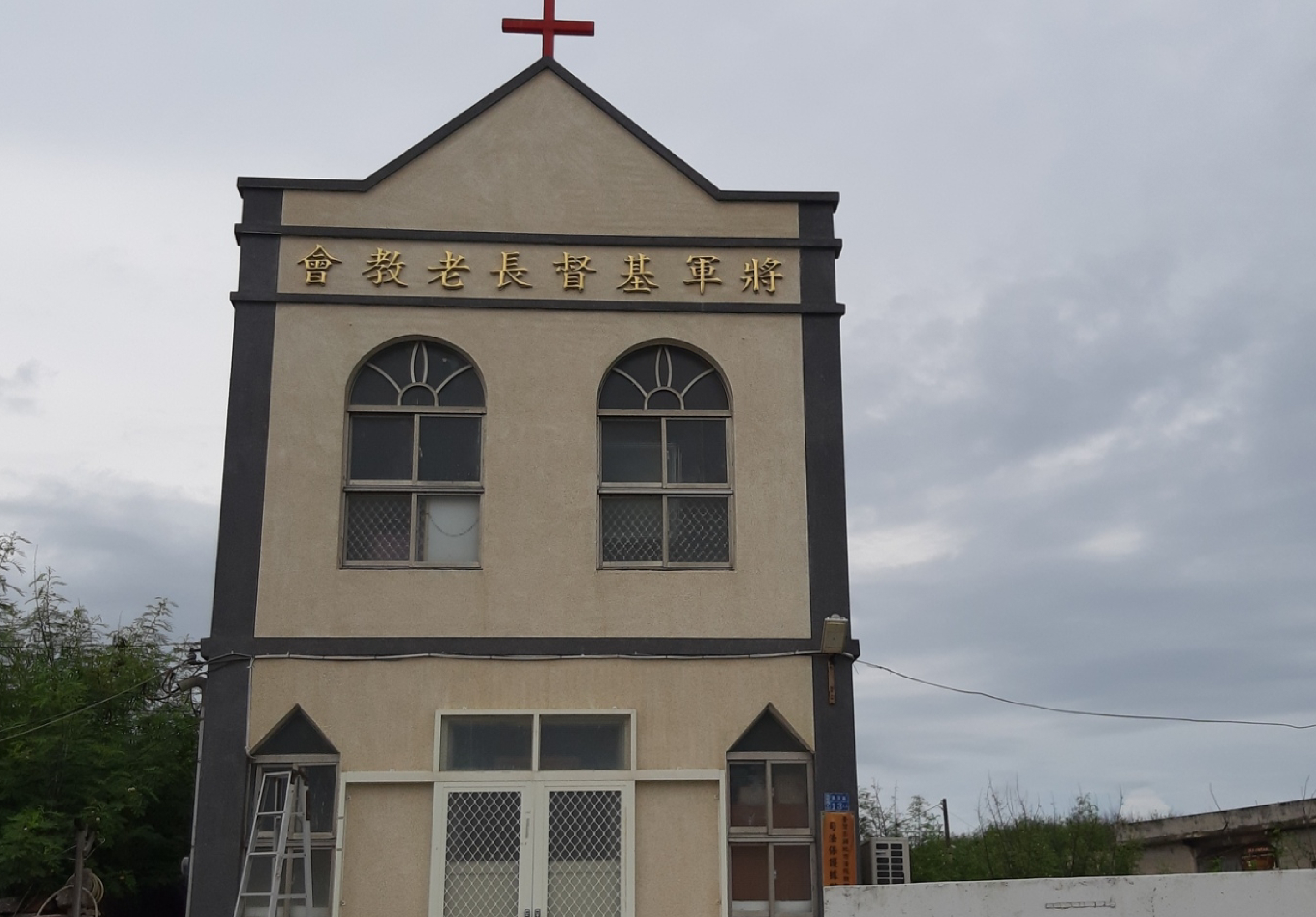 一棟米色磚造教堂建築，頂部豎立紅色十字架。建築牌匾上刻有繁體中文「會教老長晉基軍將」。正面有兩扇半圓形窗戶和下層雙扇門窗，窗戶配有網格玻璃。灰色天空背景，建築周邊可見電線和植被。