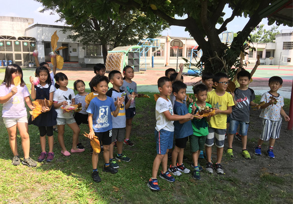 一群小學生在校園大樹下聚集，手持黃色物品，穿著夏季校服和運動服。背景是校舍和遊樂設施。孩子們似乎在進行集體活動或課外教學，呈現歡樂的校園學習氛圍。