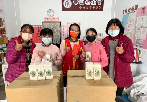 五位戴著口罩的人員在室內展示茶葉產品，站在兩個紙箱前，做出豎起大拇指的手勢。背景牆上貼著中文海報與宣傳資料，環境為社區或展示空間。