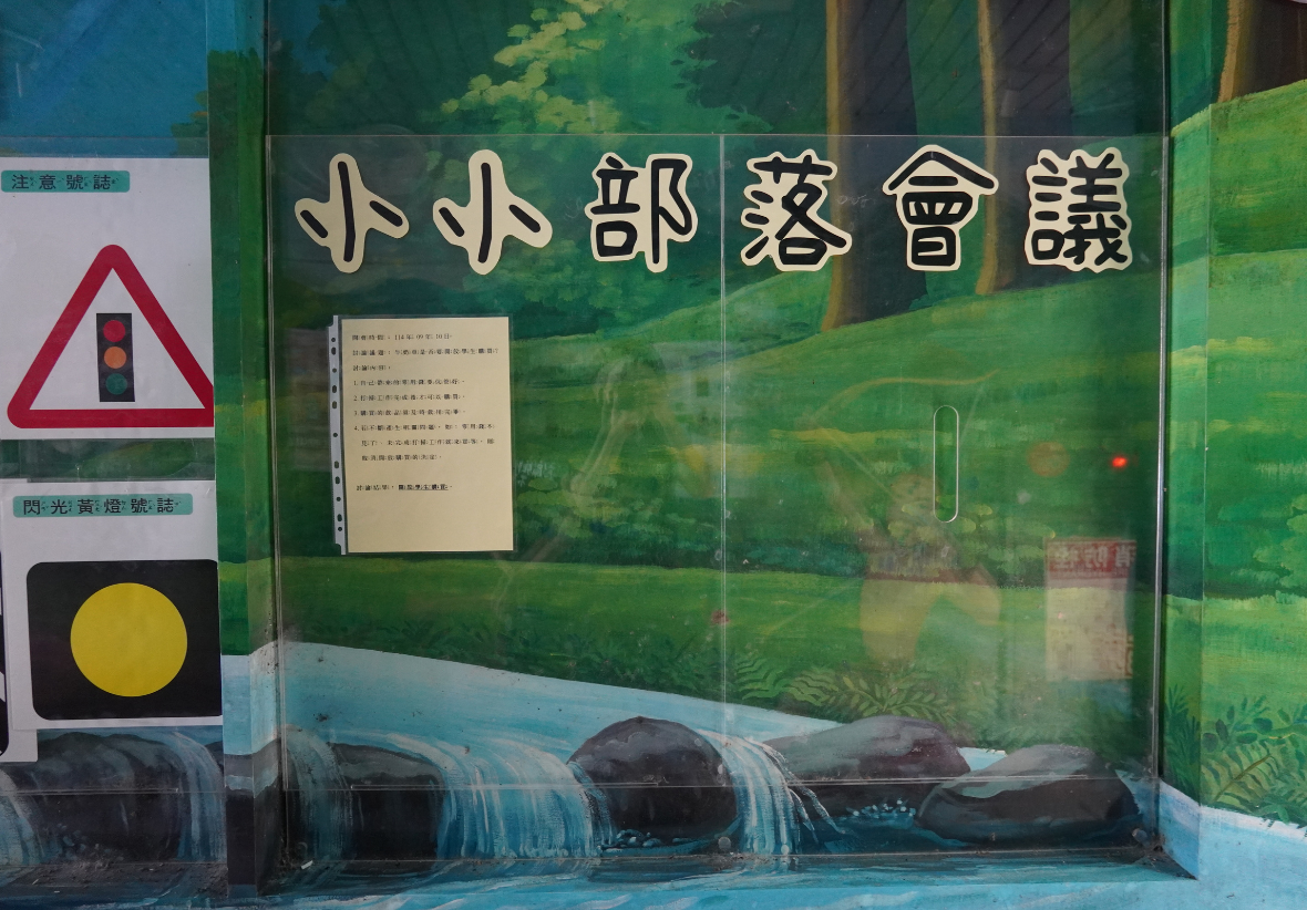 小小郵遞會議展示牌，搭配綠色自然背景和流水景觀。左側有紅色警告三角形標誌和黃色圓形信號燈。中央貼有白色說明文件。上方以金色大字標題展示，下方為藍色水景和黑色管道裝置。