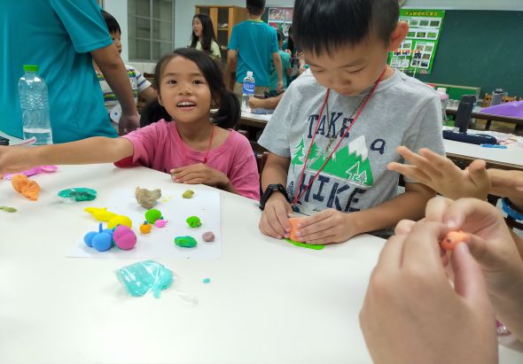 教室內，兩名小學生坐在白色桌子前進行手工藝活動，桌上擺放多彩的黏土和手工材料。女孩穿著粉紅色上衣，男孩穿著灰色上衣，兩人專注地進行創意製作。背景可見其他學生和穿著藍色制服的成人。