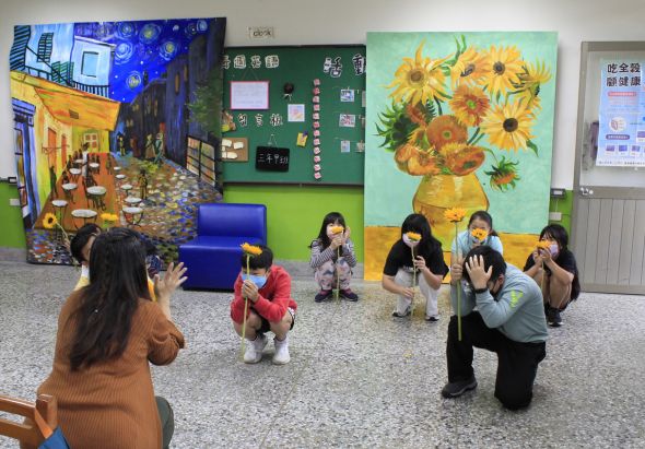 藝術教室內，一位成人在教導多位兒童進行藝術創意活動。牆面展示有梵谷向日葵畫作複製品、夜間街景藝術作品及電路板主題的互動展示，藍色坐墊置於地上。兒童蹲坐或站立於空間中，專注於學習互動。