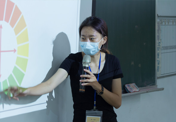 一位戴著藍色口罩的女性講者在教室內進行演講，手持麥克風，身穿黑色短袖衣服並配戴識別證。身後的投影幕上顯示一個彩色的圓形圖表，左側可見綠色黑板。