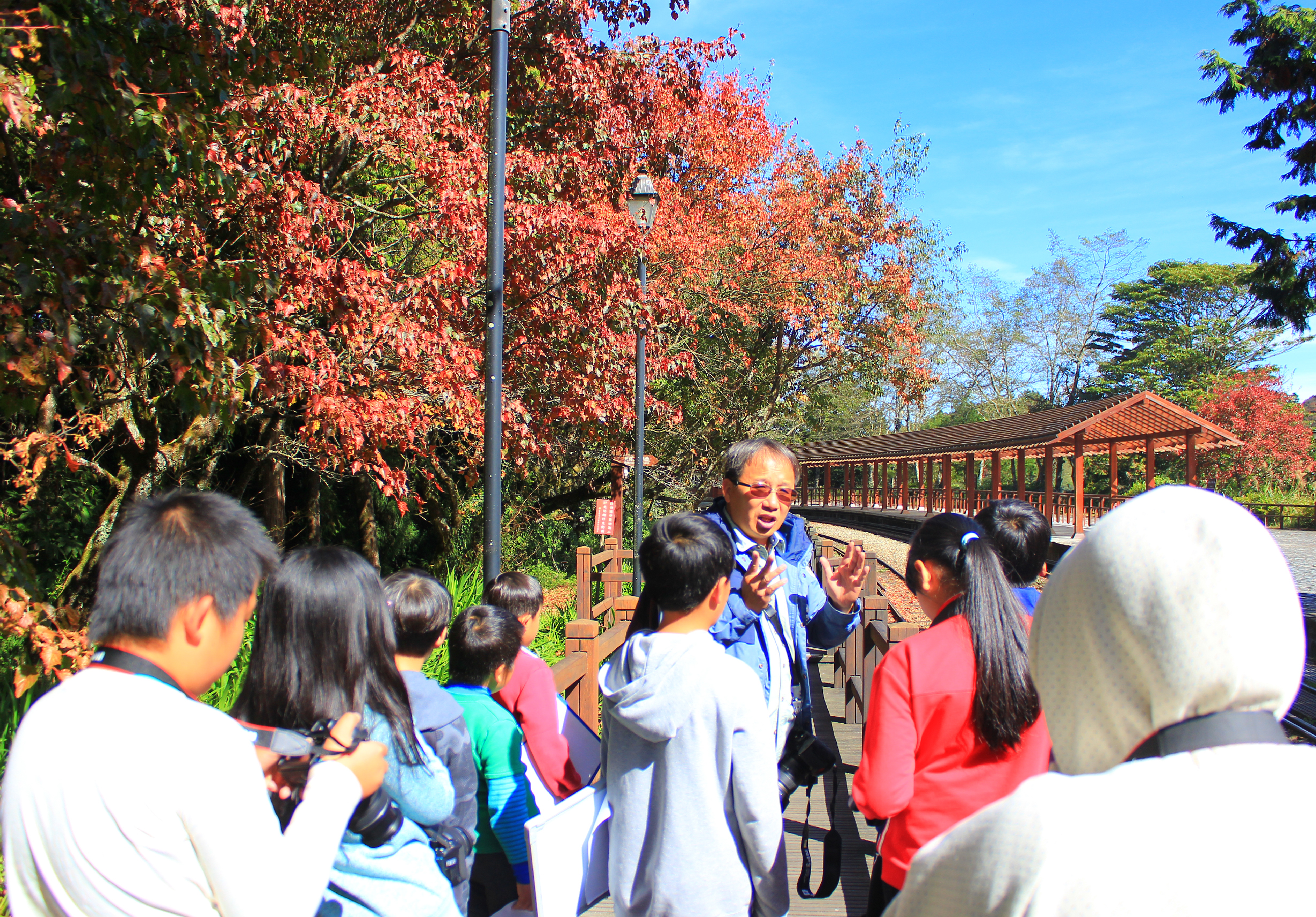 秋季公園導覽活動，一位戴眼鏡的男性講師面向遊客群眾進行解說，身後是紅色楓葉步道和木製涼亭，遠處可見變紅的樹木和晴朗藍天。