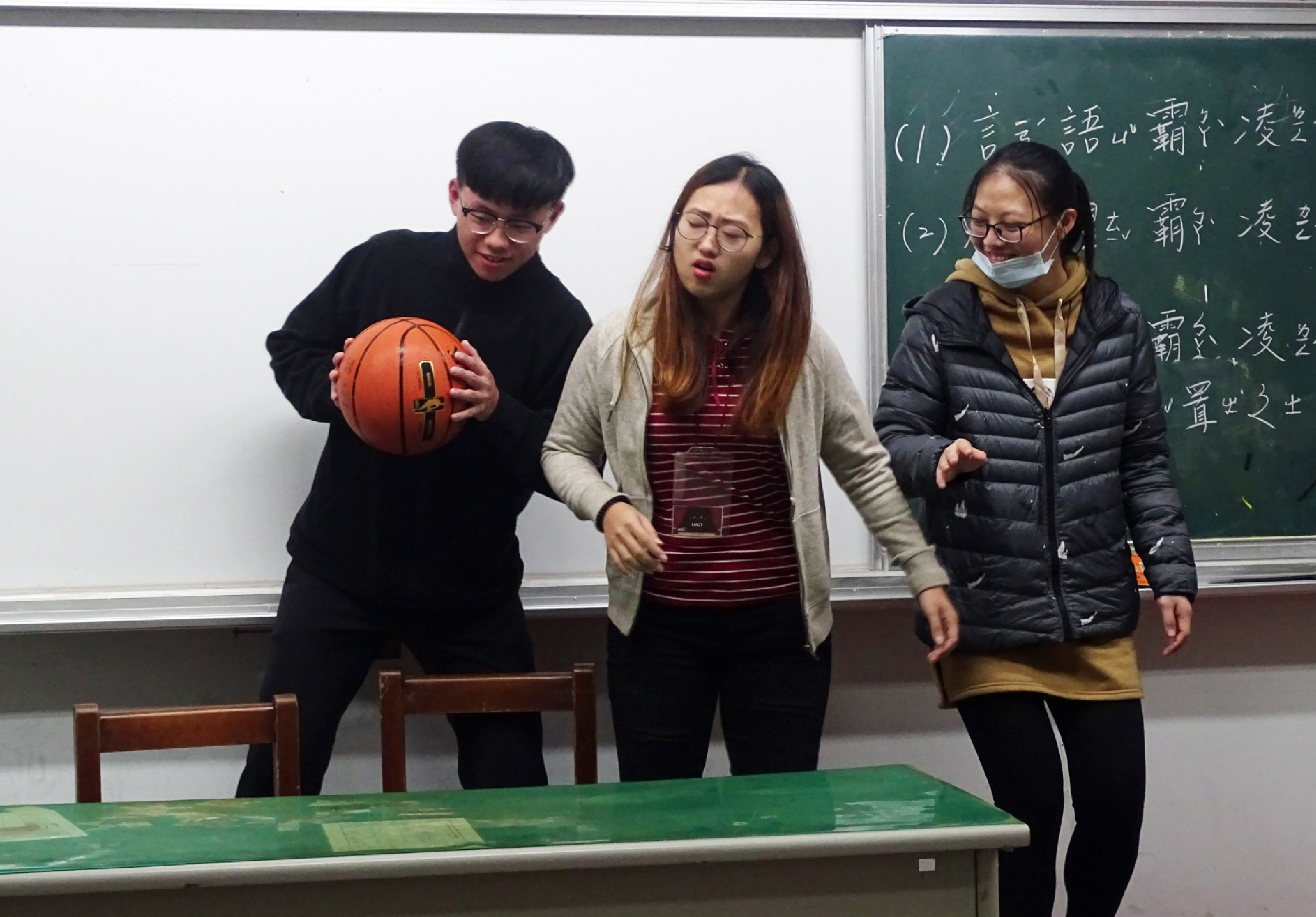 教室內三名學生站在講台前進行簡報。左方學生穿黑衣持籃球，中央學生穿紅條紋衣與米色外套，右方學生穿黑色羽絨衣並配戴口罩。身後綠色黑板寫有中文字，前方有綠色講桌。