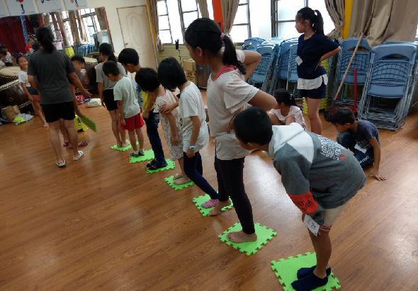 室內體育課程中，多名兒童穿著運動服站在綠色方形墊子上排成一列，跟隨成人教練進行運動訓練。木質地板教室內可見藍色椅子和教學用具，窗戶採光充足。