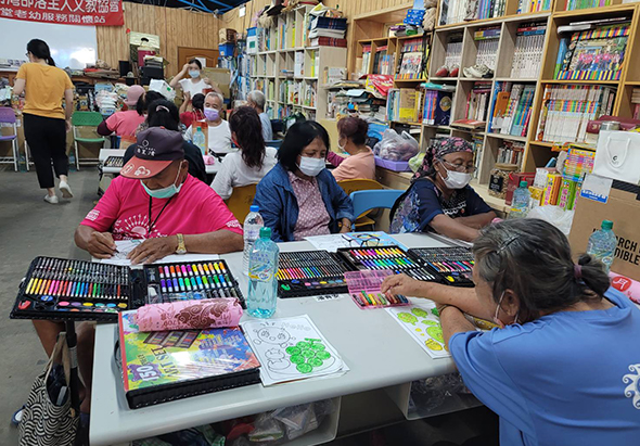 圖書館內舉辦兒童美術課程，多位小朋友坐在桌前使用彩色筆和畫具進行繪畫創作，背景有書架、其他參與者及教學人員，呈現充滿學習氛圍的活動場景。