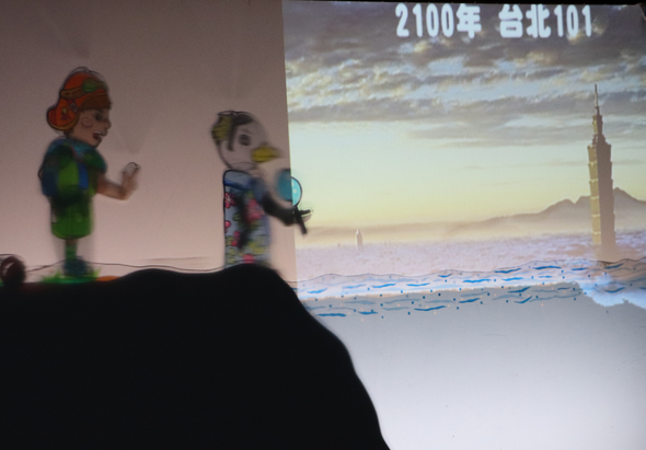舞台上兩個卡通人物角色進行表演，身後螢幕播放未來城市景象，標題顯示「2100年 台北101」，呈現台北101在未來的想像景觀，講者正向觀眾說明未來城市願景的演講場景。
