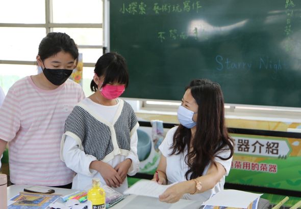 課堂中三名戴口罩的學生圍在講桌前，進行科學實驗。左側學生穿粉紅條紋衣服，中間學生戴粉紅口罩，右側學生穿白色上衣。桌上放有黃色液體瓶和實驗用具。背景為黑板，寫有中文和英文內容。