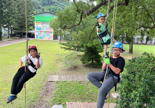 公園內三位戴著安全帽的參與者在進行高空繩索體驗活動。他們穿著安全裝備，分別在不同高度的繩索上擺出動作。綠色草地、樹木和遠方的告示牌構成背景。