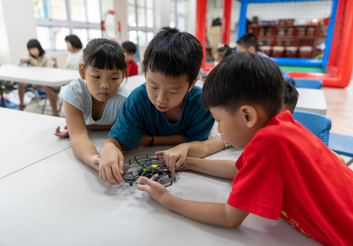 三名小學生在白色桌面上專注地組裝機器人模型，身穿不同顏色的短袖衣服。教室內可見其他學生和紅藍色充氣遊樂設施，展現STEAM教育的學習環境。