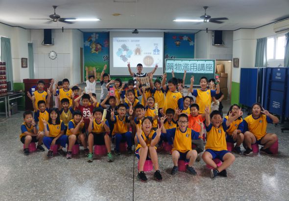 小學教室內，約五十位穿著黃色運動服的學童集合拍照。教室前方有投影幕、黑板和教學資源，學童們排成多排蹲姿或站姿，展現活力與團隊精神。