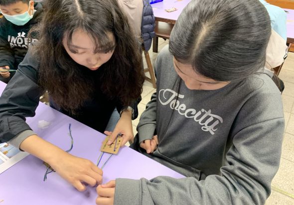 兩名女性在室內手工藝活動中，在紫色紙張上進行製作，左邊女性穿黑衣戴口罩，右邊女性穿黑色上衣專注地進行手工操作，背景可見其他參與者。