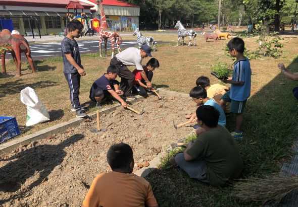 一群兒童在戶外空地上參與園藝活動，聚集在已翻鬆的土壤周圍，成人指導他們使用工具挖土和種植。背景可見遊樂設施和樹木，陽光灑落在活動場地上。