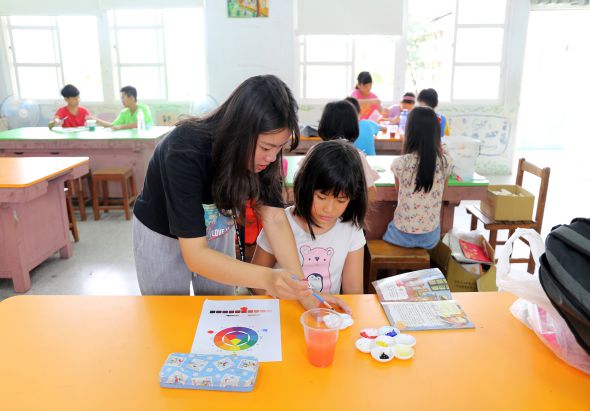 幼兒園教室內，一位女教師與小女孩在黃色桌前進行美術活動，桌上擺放著彩色輪盤、畫紙等教材。背景有多位幼童在教室各處參與學習活動，明亮的教室窗戶採光充足。
