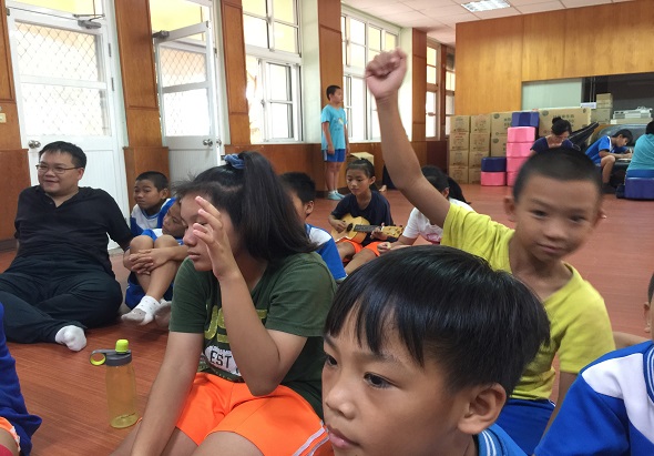室內體育館中，多名穿著彩色運動服的兒童坐在木製地板上參與集體活動。前景兒童舉起手臂，背景有成人及其他兒童。牆面為木質裝飾，窗戶採光充足，呈現熱鬧的兒童運動課程或團隊活動場景。