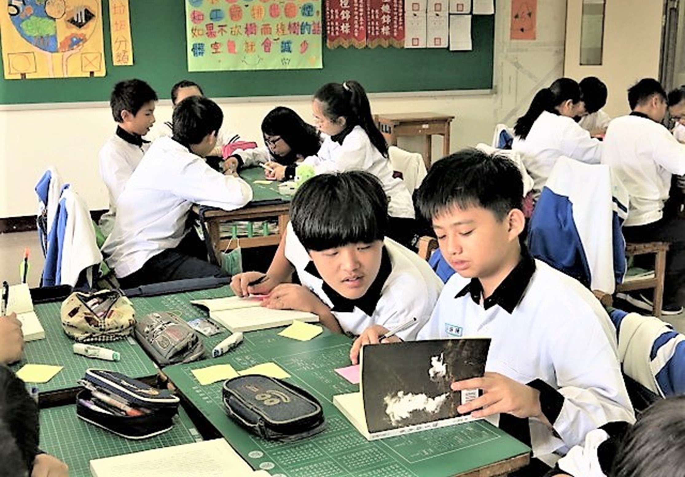 教室內，穿著白色制服的學生坐在綠色課桌前進行美術課程。前景兩名學生正在查看黑色素描本，背景有多位學生進行各種創意活動。牆上貼著彩色教學海報和中文標語。