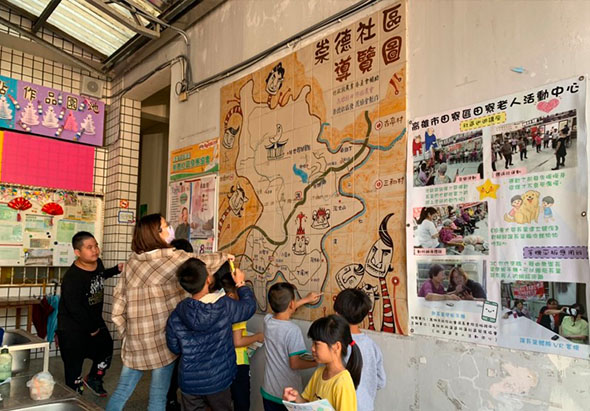室內教室場景，牆上貼著手繪地圖和活動海報。五位兒童圍著大型棕色地圖板互動，有大人在旁指導。牆面右側展示多張彩色照片和文字資訊。左上方有電視螢幕播放內容。此為兒童教育活動或導覽學習情景。