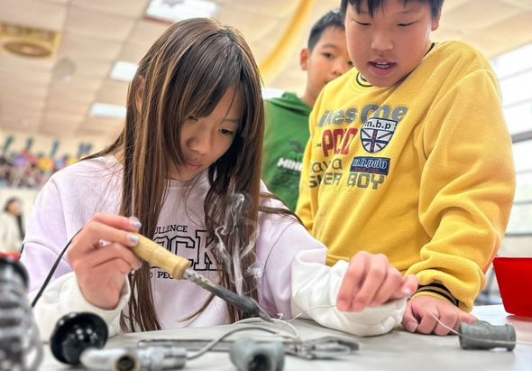 室內教室中，穿著白色和黃色衣服的兒童聚集在桌邊，專注地進行科學實驗或手工活動。他們正用工具檢查或組裝某個物品，展現了學生在課堂上的互動學習情景。