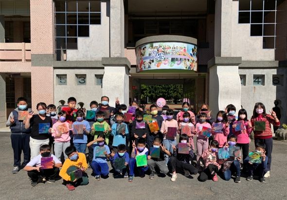 幼兒園戶外集合照，約五十位穿著彩色衣服的幼童和教職員站立在校舍前，背後有圓形彩繪建築物和綠色植栽，全體面向鏡頭擺pose拍照。