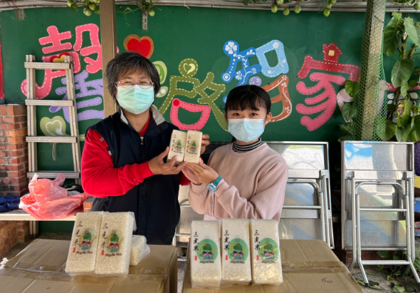 兩位戴口罩的年輕人在綠色背景的展示區前展示米製產品。桌上擺放多個包裝精美的白色米袋，背景牆面裝飾著繽紛的紅藍黃色中文字及節日裝飾。左側展示架上放置花卉擺設，呈現農產品推廣活動氛圍。