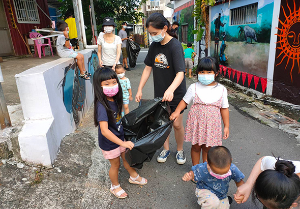 戶外街道上，一位成人與多名小孩一起進行垃圾分類活動。他們手持黑色垃圾袋，正在清理社區環保。背景可見住宅建築和彩繪牆面。參與者都戴著口罩，展示社區環保教育的實踐。