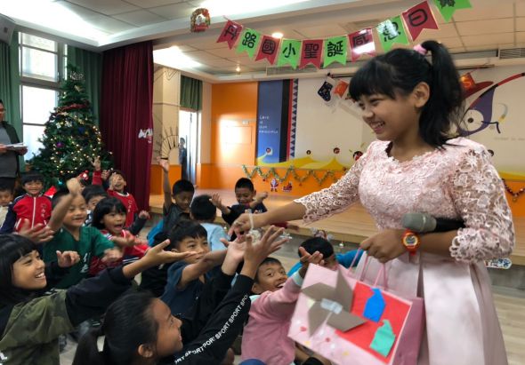 小學教室內，一位女教師穿著粉色蕾絲上衣，正向圍坐在她身邊的學生們發放或展示小國旗。教室內裝飾有聖誕樹、彩色紙旗和國旗，多名學生穿著校服認真聆聽。