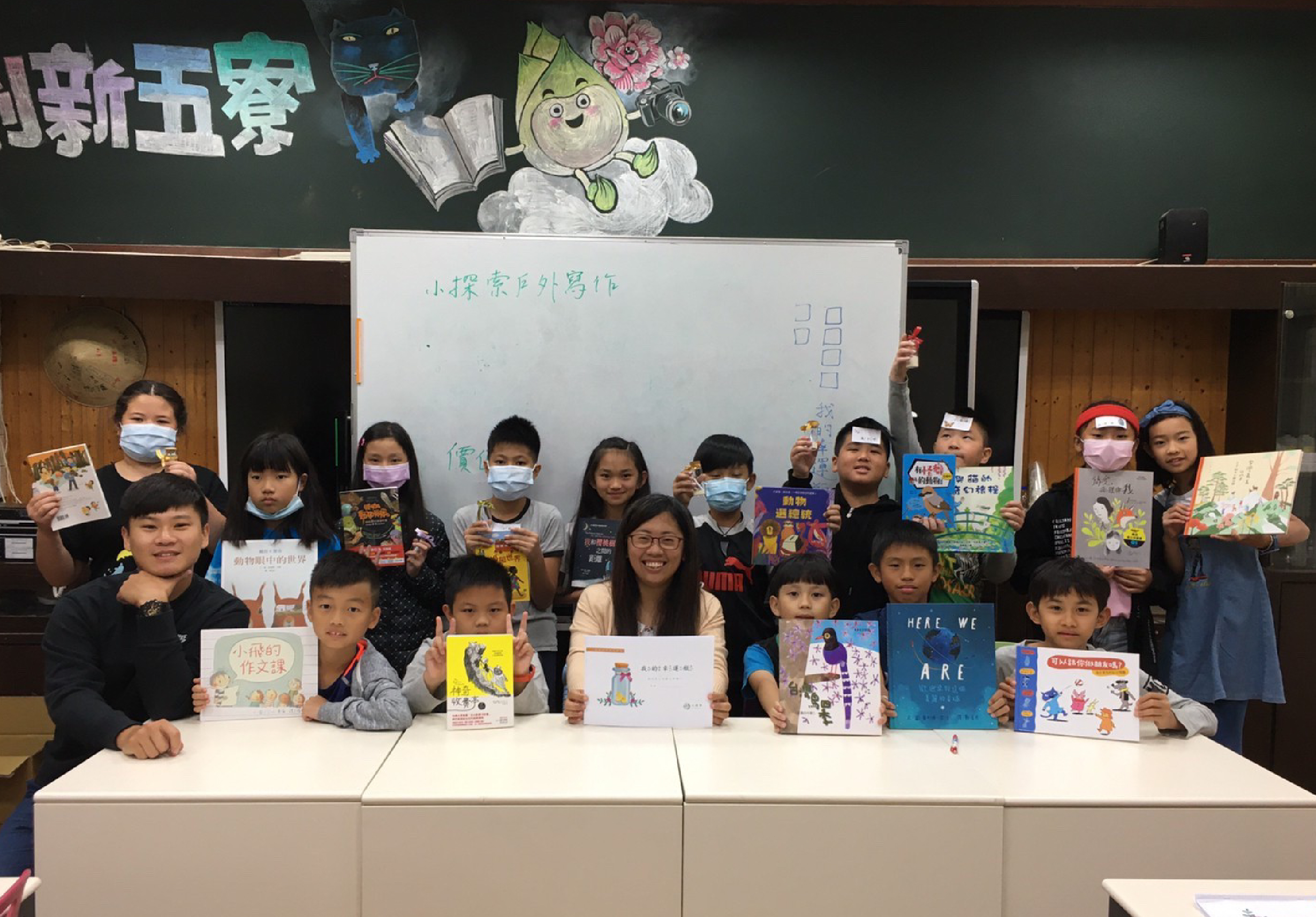 小學教室內，一位女教師與約二十位學生在講臺前合照，學生們手持各式兒童書籍和作品展示。黑板上寫著「小探索百外寫作」，背景有卡通角色裝飾。多數學生戴著口罩，展現閱讀與寫作學習成果。