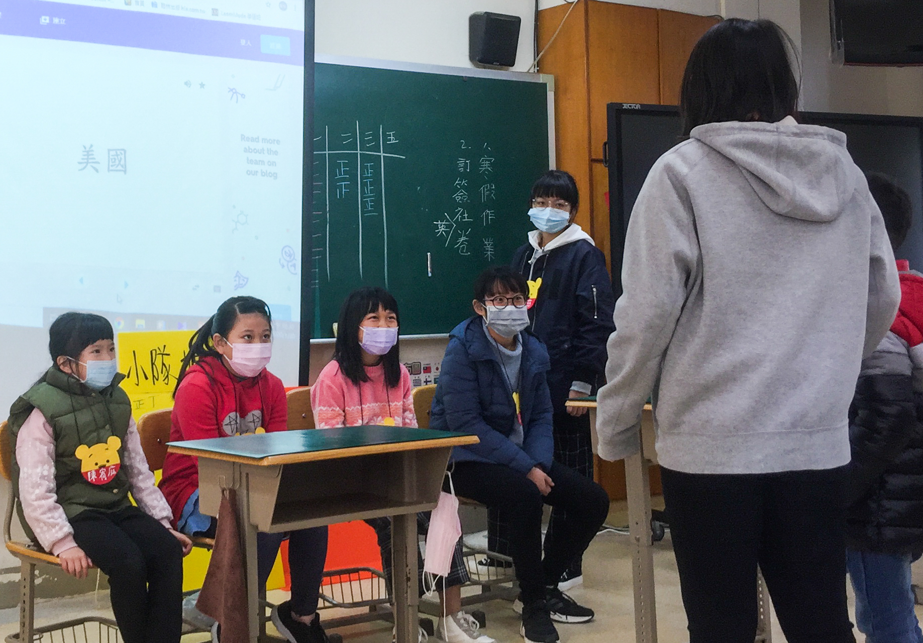 教室裡進行課堂活動，多名配戴口罩的學童坐在課桌前聽講，講台上有黑板寫著中文字，牆上投影螢幕顯示教學內容，一名站立的學生舉手回答問題，教室後方可見教師身影。