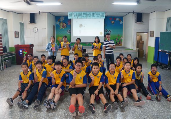 小學教室內，約二十名穿著黃藍色運動服的學生坐成一圈，五位老師站在後方。教室前方有藍色布置的教學牆面和投影螢幕，場景顯示為班級體育或團隊活動的合照。