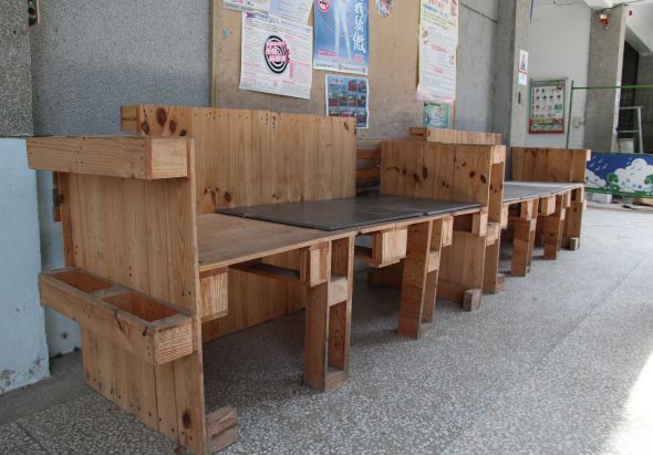 室內空間展示一套木製餐桌椅組合，採用淺色木材製作，桌面為深色材質。桌邊配置多張高背木椅。背景牆面貼有各類公告和海報。地面為淺色地磚。