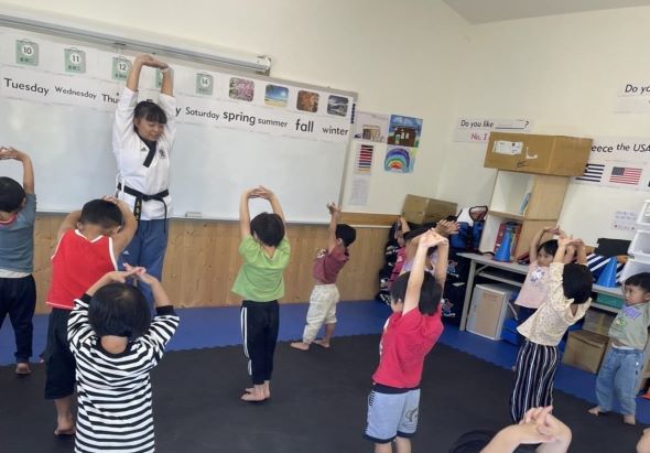 教室內，一位穿著白色道服的武術教練正在教導約十名幼童練習武術動作。孩童們穿著不同顏色衣服，站在藍色墊子上，跟隨教練做出舉手等基本招式。牆上貼有日期和季節標籤。