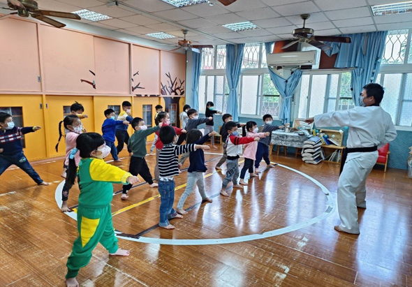 學校體育館內，一位教師指導約二十名小學生進行運動課程。學生們穿著不同顏色的運動服，在木質地板上排成圓形，參與團體體操或舞蹈活動。教室牆面為粉紅色和黃色，窗戶採光充足。