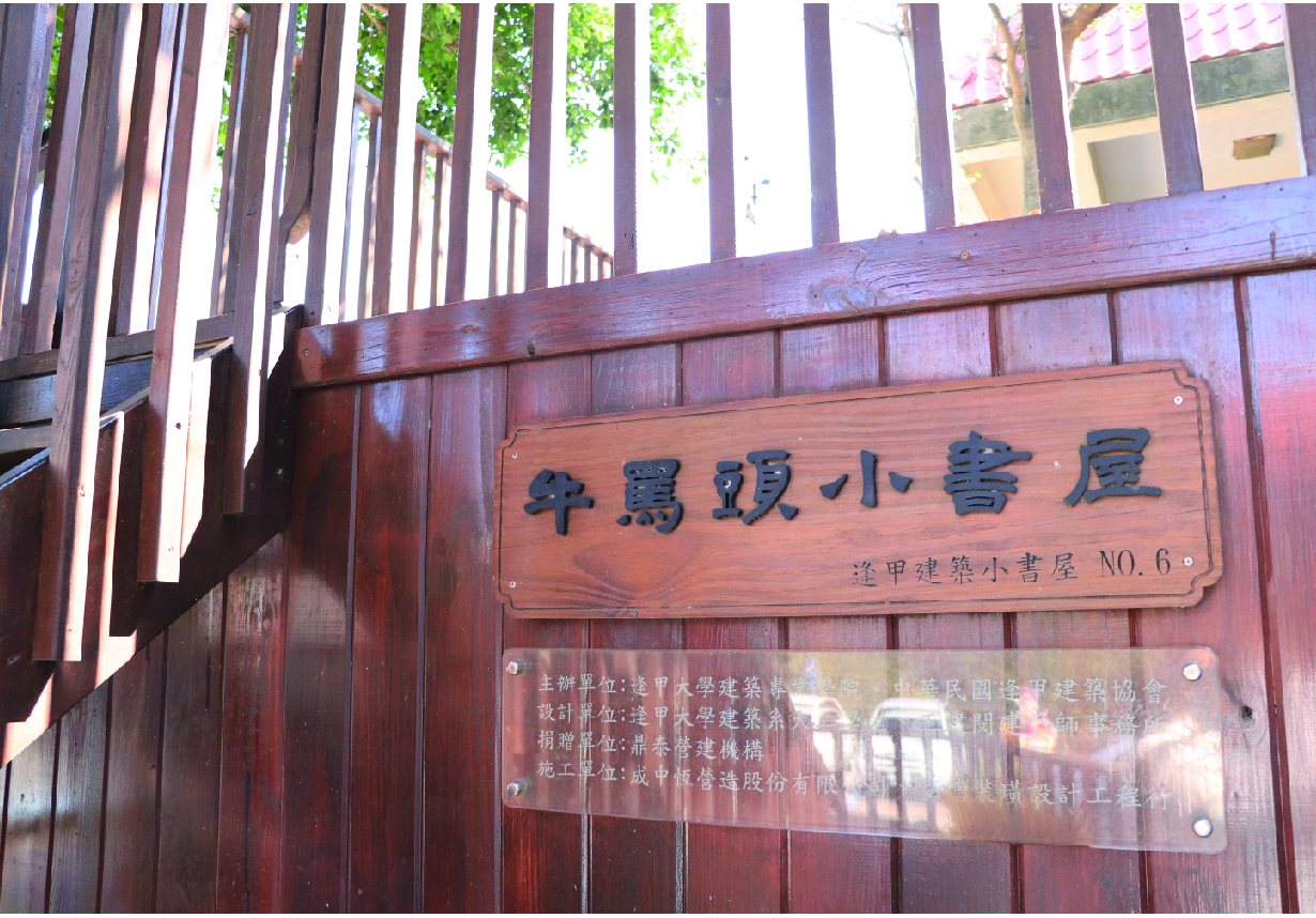 紅褐色木製建築外牆上掛著木製招牌，上方刻有繁體中文「牛窩頸小書屋」，下方標註「港甲建築小書屋NO.6」，招牌下方附有說明銘牌。建築左側為豎條木柵欄，上方可見綠色植物和建築物。