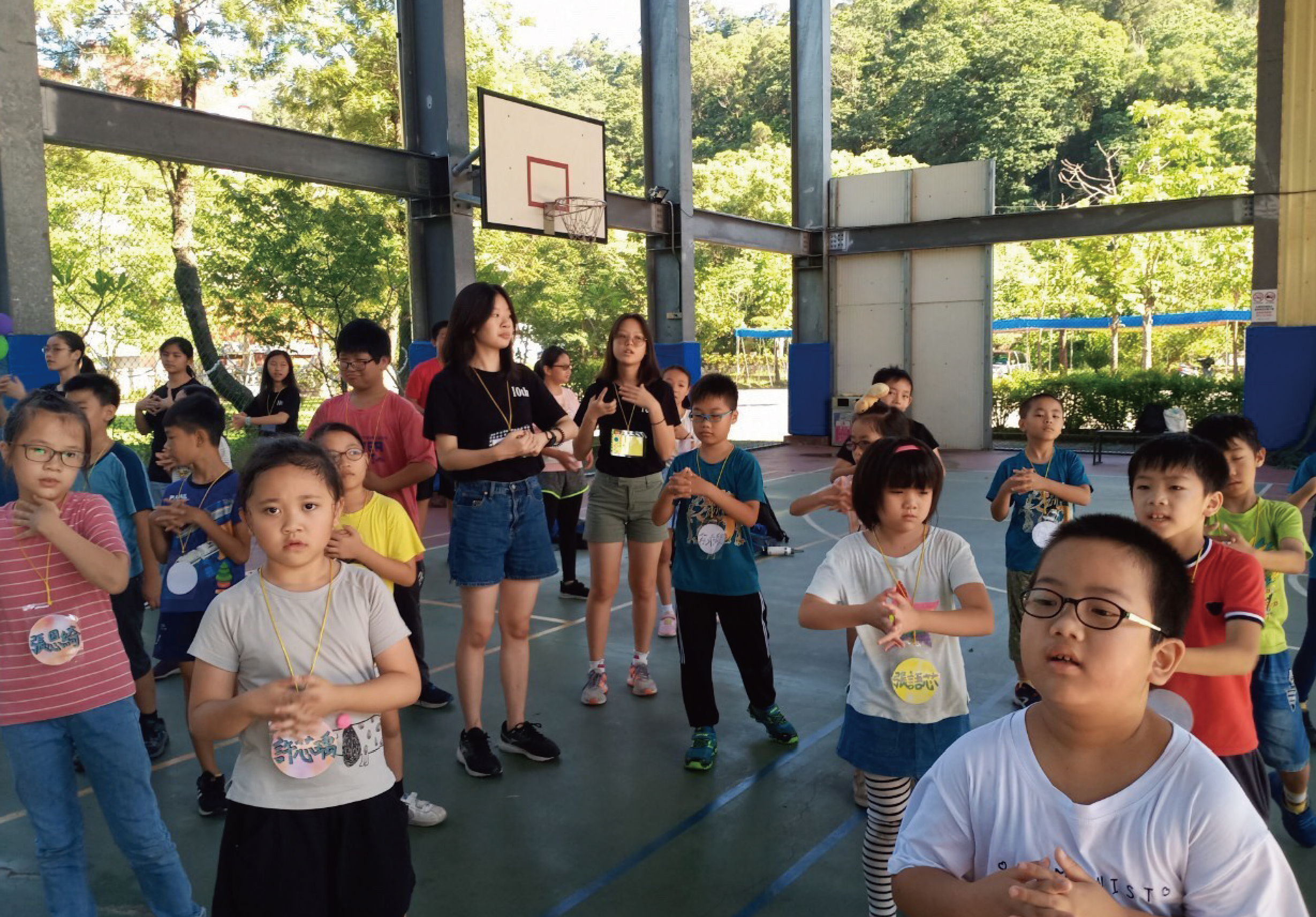 室內籃球場進行兒童運動課程，約二十多位小學生穿著運動服，跟隨女性教練做伸展運動。背景可見籃球架、綠色樹木與藍色圍欄，學生們認真參與體育活動。