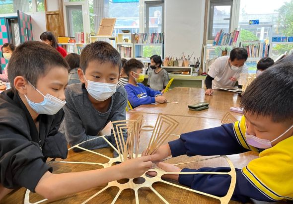 小學生在教室內進行木工藝術活動，戴著口罩圍坐在木製工作台周圍，合作組裝木製星形結構。背景可見書櫃、窗戶和其他學生在進行創意勞作。