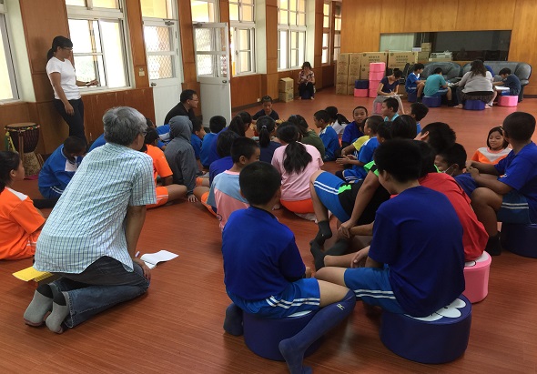 室內教室，一名年長男子與眾多穿著藍色和橙色服裝的兒童圍坐成圈，進行互動式團體活動。教室裡有木質牆壁、黑板和粉紅色塑膠籃子，營造出學習和社區參與的氛圍。