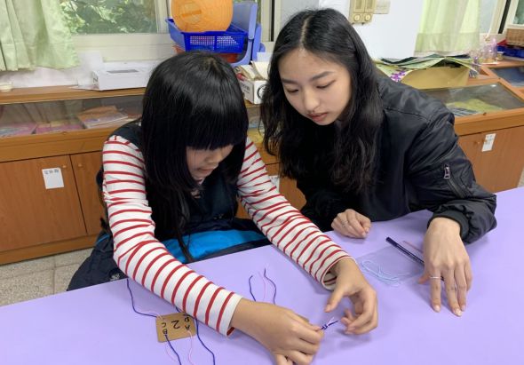 兩位女學生在教室紫色桌面上進行美勞課程。左邊學生穿著紅白條紋長袖衣著，右邊學生穿著深色外套，兩人專注地在紙上繪畫創作。