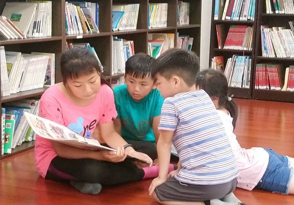 三名小學生坐在圖書館地板上，聚集在一起閱讀一本繪本。穿著粉紅色衣服的女孩持書，其他兩位男孩專注地看著書頁。身後是擺滿各類書籍的木製書架。