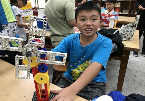 一名穿著藍色T恤的男童在教室內展示自己製作的彩色機械裝置，該裝置由樂高積木和各色輪片組成。背景中可見其他學生和成人正在進行科學或工程相關的動手活動。