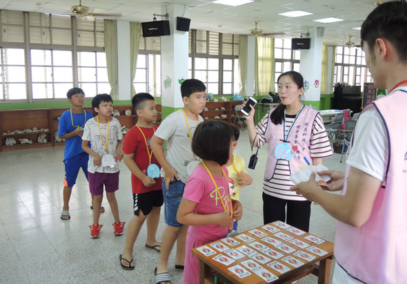 一群小學生在寬敞的室內場地參與教育活動，老師和志工正在進行互動式教學。地上擺放著裝有學習卡片的盒子，學生們穿著彩色衣服專注參與，展示出活潑的課堂學習氛圍。