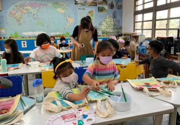 教室內，小學生們戴著口罩進行美術課程。學生坐在白色課桌前，用顏料和筆在紙張上創作。教師站在後方指導。牆上貼著世界地圖和教室佈置。學生認真投入於繪畫活動中。