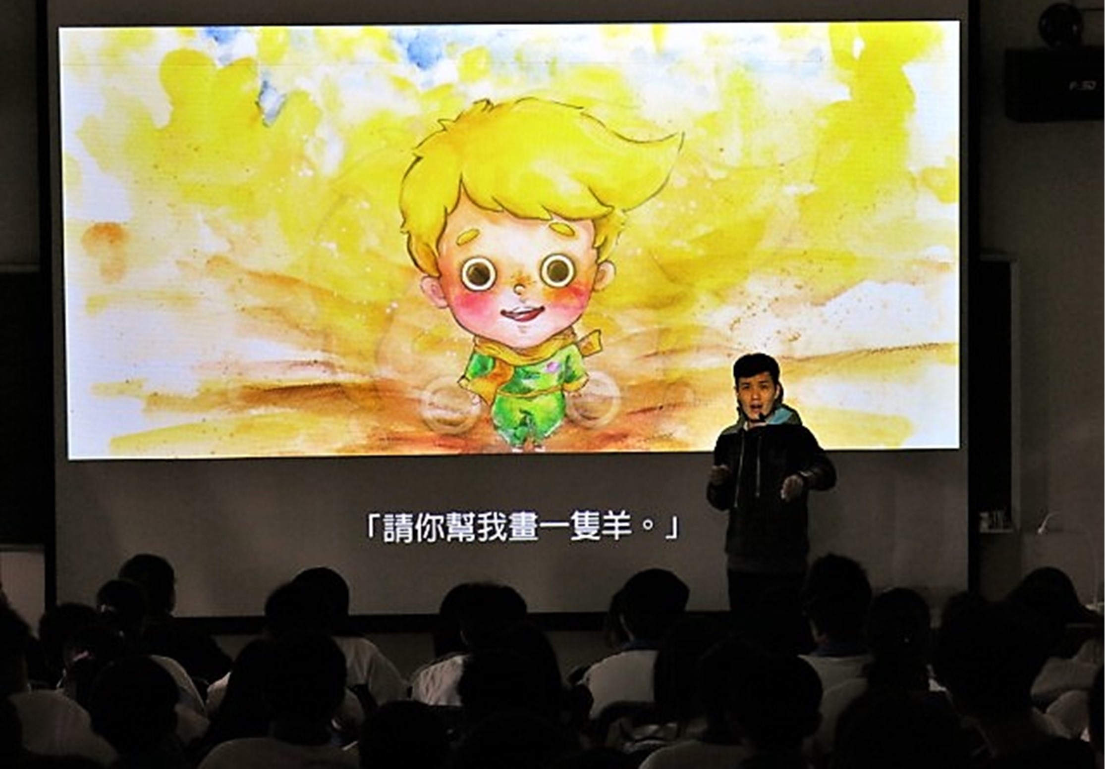 講者站在舞台上，身後投影螢幕顯示一個黃髮、綠衣服的卡通小男孩角色，背景為黃色光暈。螢幕下方有繁體中文字幕「請你幫我畫一隻羊。」觀眾坐在黑暗中觀看演講。