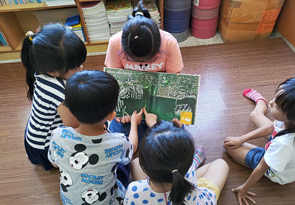 一位成人老師坐在地板上，與五名幼童圍坐一圈，共同查看一本綠色封面的圖書。室內背景有書架、儲物籃和教室用品，呈現溫馨的集體閱讀教學場景。