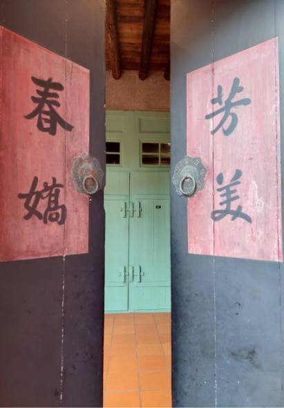 傳統建築入口，綠色木門兩側各有一幅紅色對聯，左側書寫「春嬸」，右側書寫「芳美」，門上方有木製橫樑，下方為橙色磁磚，展現古典東方建築風格。
