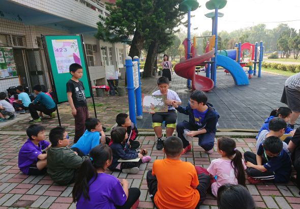 學校操場上，一群穿著彩色運動服的小學生圍坐成圈，正在參加戶外教學活動。中央站著帶領活動的老師，背景可見紅藍色溜滑梯遊樂設施、綠樹和校舍建築。